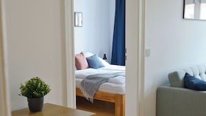 Una camera da letto moderna con pavimento in legno, un letto con cuscini blu e rosa, un divano grigio e un piccolo tavolino con una pianta.