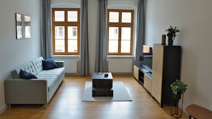 Un soggiorno moderno con un divano grigio, un tavolino da caffè e un mobile TV. Due finestre con tende, un termosifone e una pianta sul mobile TV.