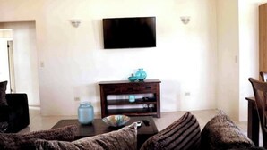 A living room with a flat-screen TV mounted on the wall, a dark wooden coffee table, and a patterned sofa. - Island gem.  Close to beach and downtown  (Saint John's)