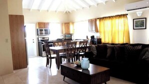 A spacious living area with a dark brown sofa, a wooden coffee table, and a dining table with chairs. A kitchen with wooden cabinets and a microwave is visible in the background. - Island gem.  Close to beach and downtown  (Saint John's)