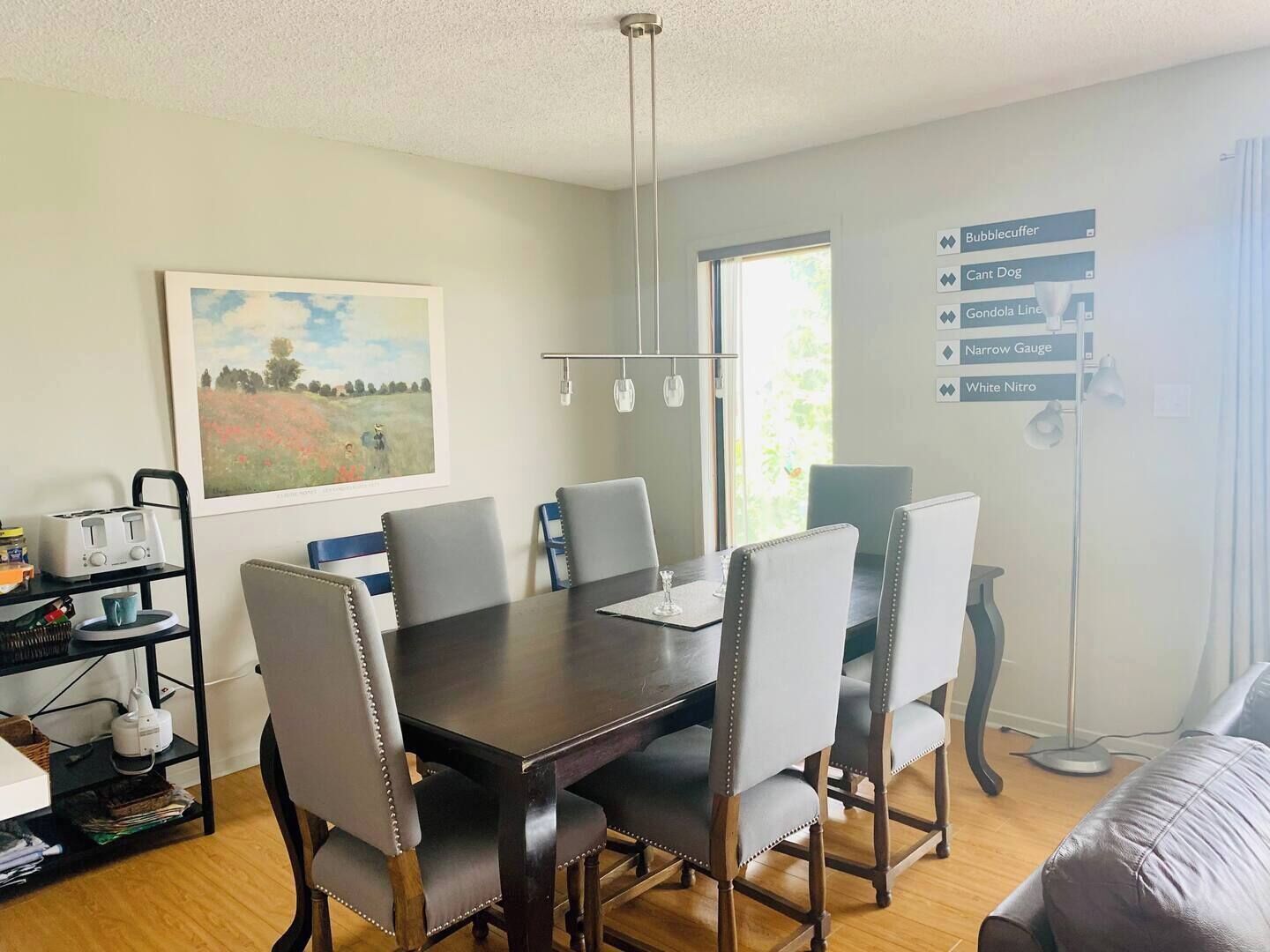 A dining area with a dark wooden table, six grey upholstered chairs, a painting on the wall, a window with natural light, and a wall-mounted light fixture.