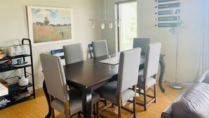 A dining area with a dark wooden table, six grey upholstered chairs, a painting on the wall, a window with natural light, and a wall-mounted light fixture.