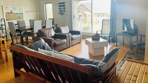 A spacious living room with a large window, a wooden sofa, a dining table with chairs, a TV, and a computer desk.