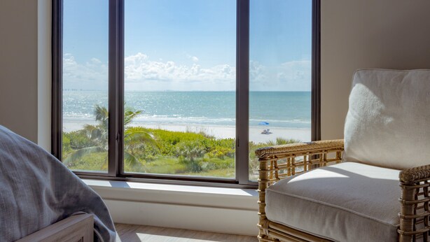 A room with a large window overlooking a beach, a wicker chair, and a view of the ocean. - All New! Gulf Front! High End! Pelicans Roost 303 on Sanibel Island (Sanibel)