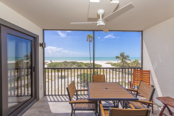 A balcony with a table and chairs, a ceiling fan, and a view of the beach and ocean.