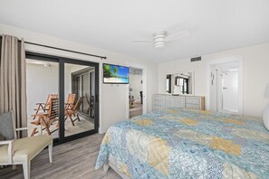 A bedroom with a large bed, a sliding glass door leading to a balcony, a ceiling fan, and a television mounted on the wall.