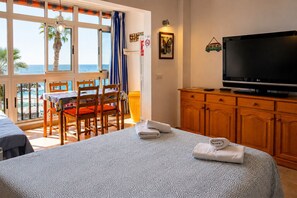 A hotel room with a bed, a television, a dining table with chairs, and a view of the sea.