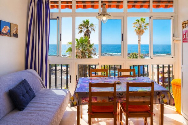 A room with a balcony, a table with chairs, a sofa, and a view of the sea.