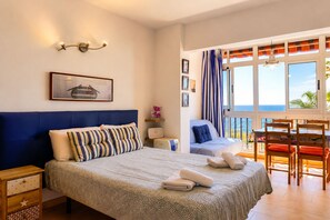 A bedroom with a bed, bedside tables, a desk, a chair, and a view of the ocean.