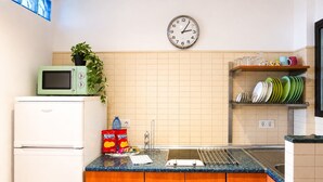 A kitchen with a microwave, a clock, a sink, and a shelf with plates.