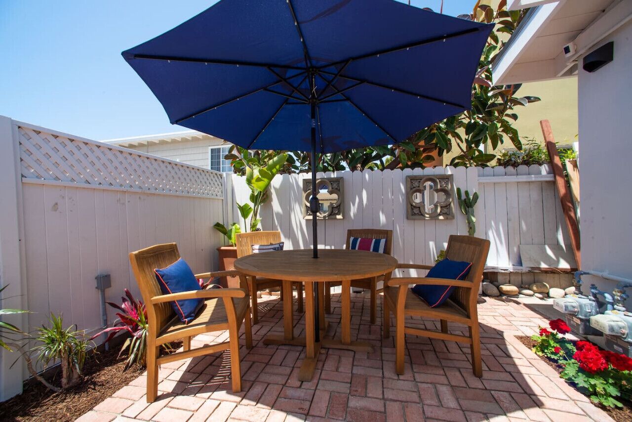 A patio with a round wooden table and chairs, a blue umbrella, and a white fence.