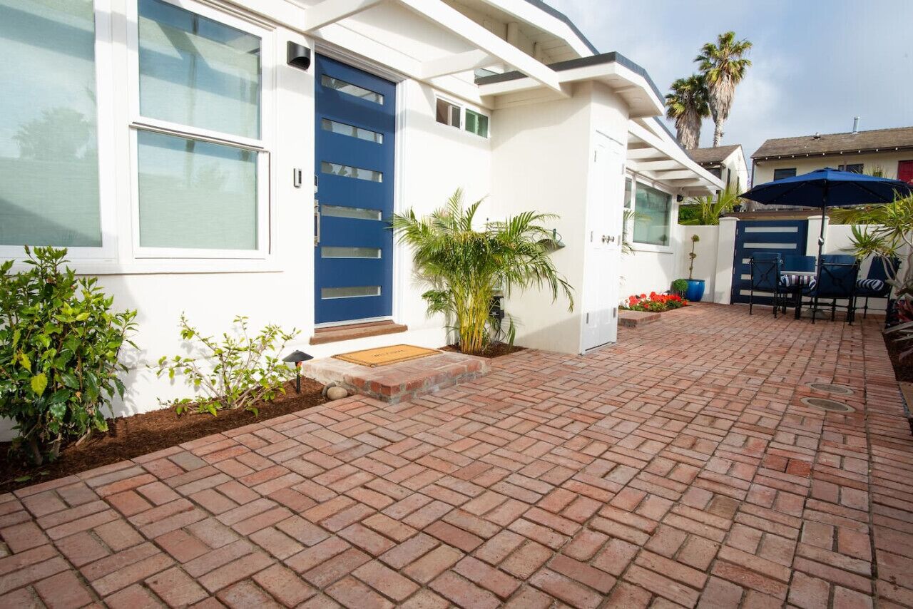 Uma casa com porta azul, um pátio de tijolos e um pequeno jardim com plantas e uma cadeira.