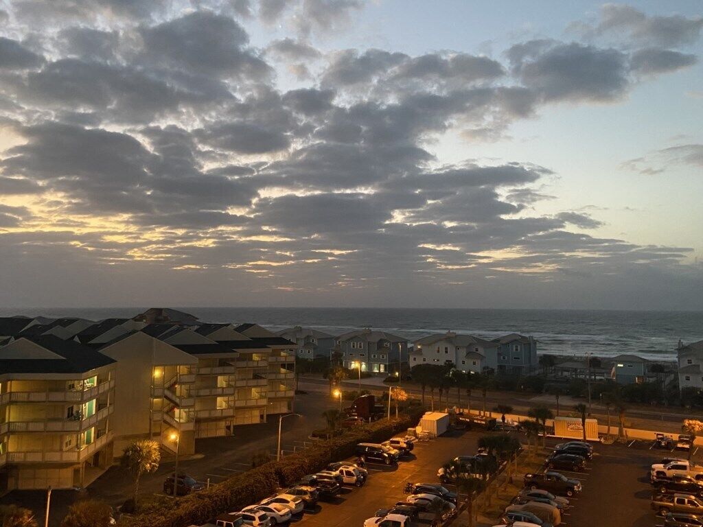 A coastal hotel with multiple-story buildings, a parking lot, and a view of the ocean.