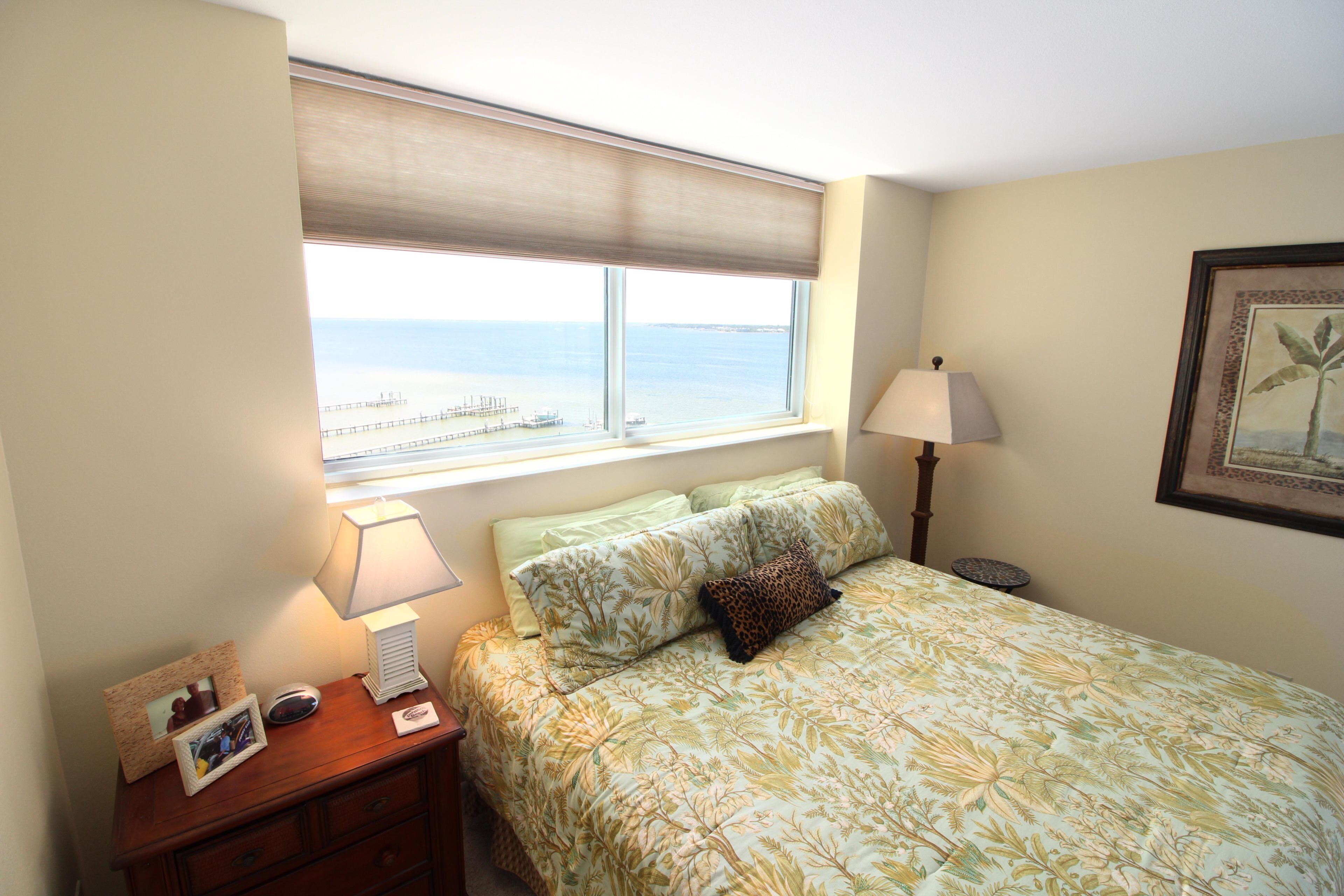 Une chambre à coucher comprenant un lit, une table de chevet, une lampe, un cadre photo et une fenêtre donnant sur la mer.