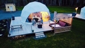 A dome-shaped tent with a wooden deck, seating area, and a small building in the background. - Beachcomber Dome Accommodations (Monks Head)