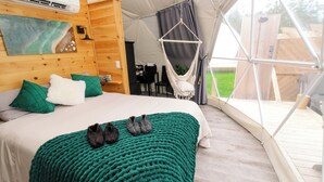A tented accommodation with a bed, green and white pillows, a green knitted blanket, and a hanging chair. - Beachcomber Dome Accommodations (Monks Head)