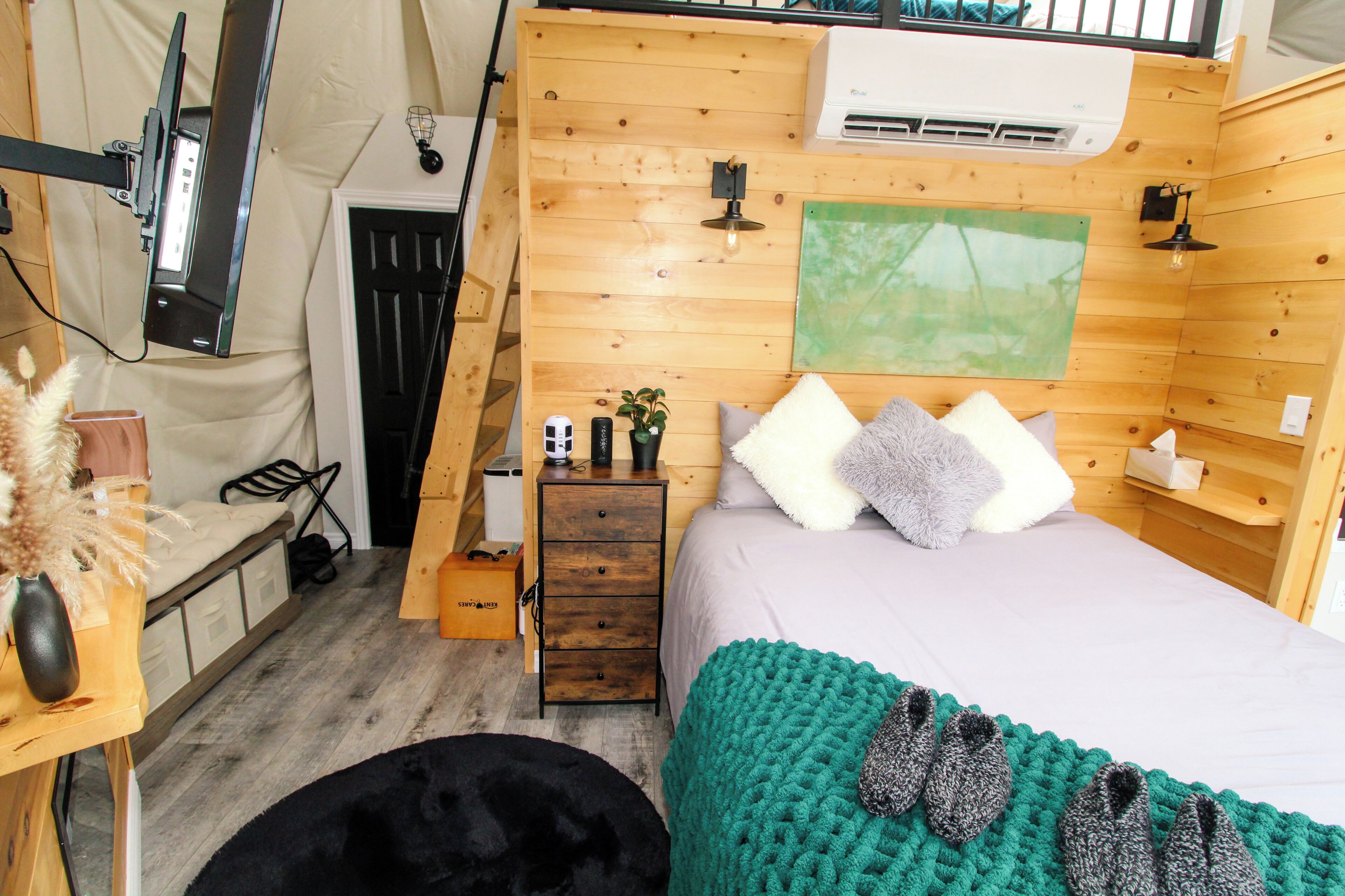 A cozy room with a bed, a small dresser, a desk with a lamp, and a wall-mounted air conditioner.