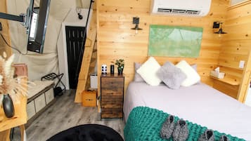 A cozy room with a bed, a small dresser, a desk with a lamp, and a wall-mounted air conditioner.