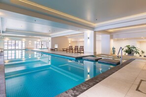 A large indoor swimming pool with a clear blue water surface, surrounded by a tiled deck area with a few chairs and a small table. - Level 25 1 Bed with Marble Lined Bath City Views (Auckland)