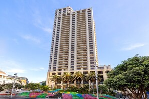 Um hotel alto, com vários andares, design moderno, com varandas em cada andar e um pátio central com palmeiras.