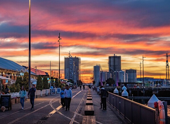 A waterfront promenade with a sunset view, industrial buildings, and a pedestrian walkway.