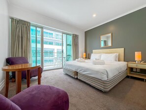 A hotel room with a large bed, a small table, a chair, and a balcony with a view of a building.