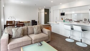 A modern living room with a sofa, a coffee table, and a dining area with chairs and a table. There is a kitchen area with white cabinets and a bar counter.