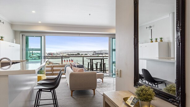 A modern living room with a large window overlooking a harbor, a kitchen area with a bar counter, and a dining space with a view. - Gorgeous Waterfront Apartment Harbour and Views (Auckland)