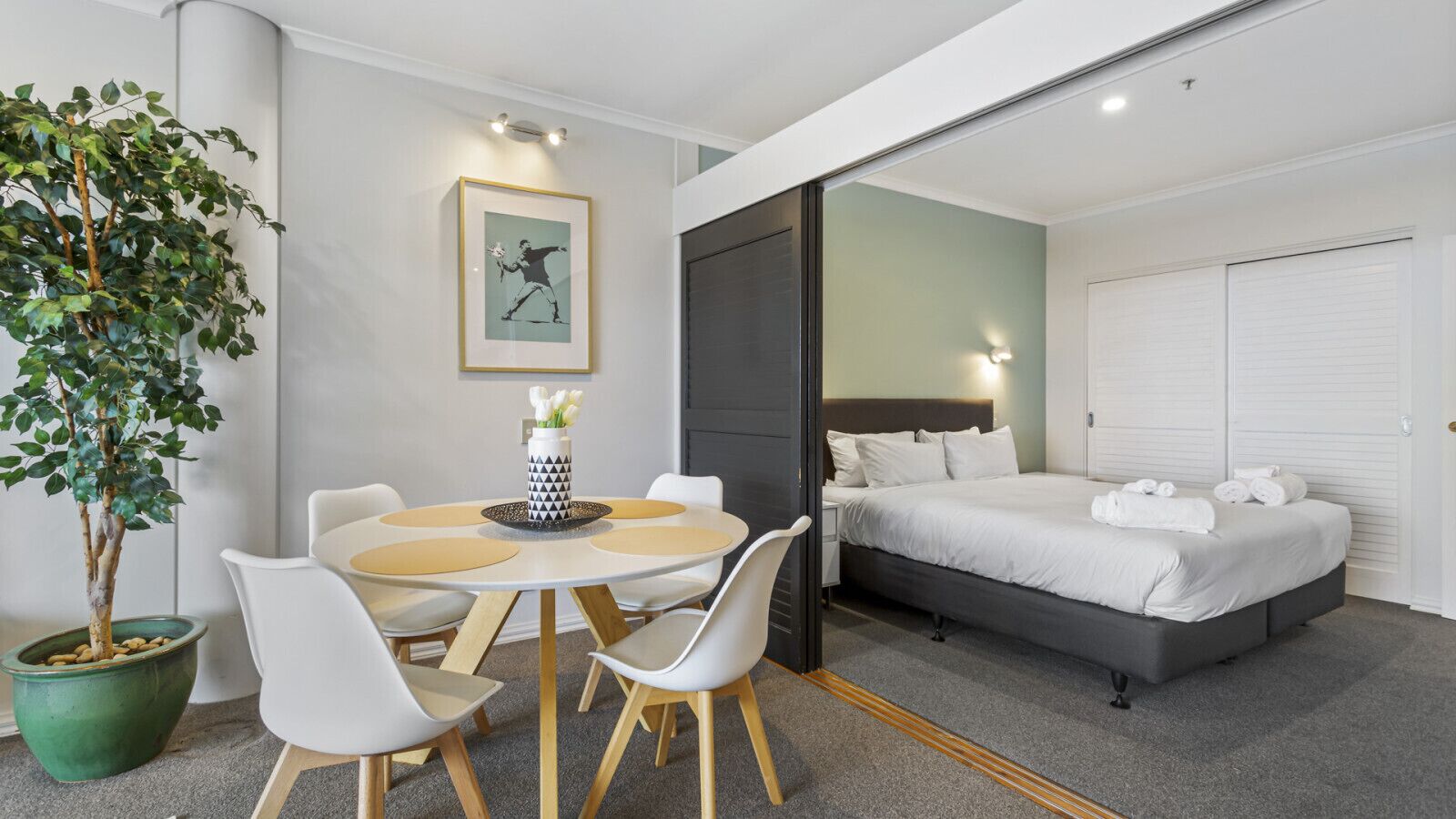 A modern hotel room with a bed, a dining area with a table and chairs, a potted plant, and a framed picture on the wall.