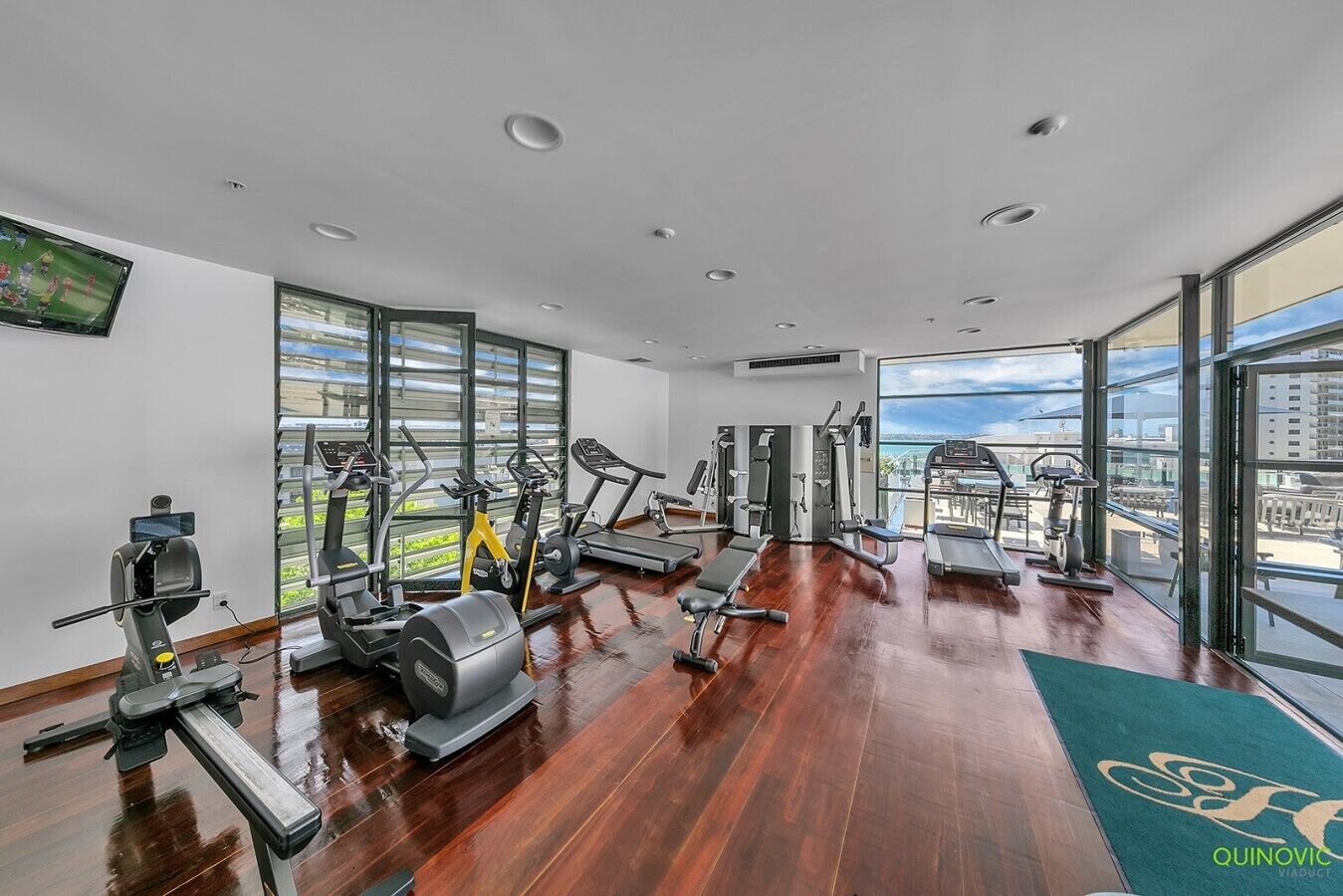 Une salle de sport équipée de divers appareils d’exercice, dotée de grandes fenêtres et offrant une vue sur la ville.