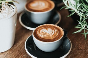 Zwei Kaffeetassen mit Latte-Art auf einem Holztisch, begleitet von einem Blumentopf und einem weißen Behälter mit weißen Perlen.