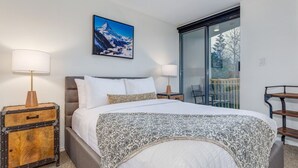 A modern bedroom with a large bed, two bedside tables, a painting of a mountain, and a balcony with a chair. - The Mountain View Condo - Large 2BR/2BA (Whistler)