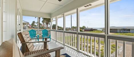 A covered porch with wooden furniture, a ceiling fan, and large windows offering a view of the beach and ocean.