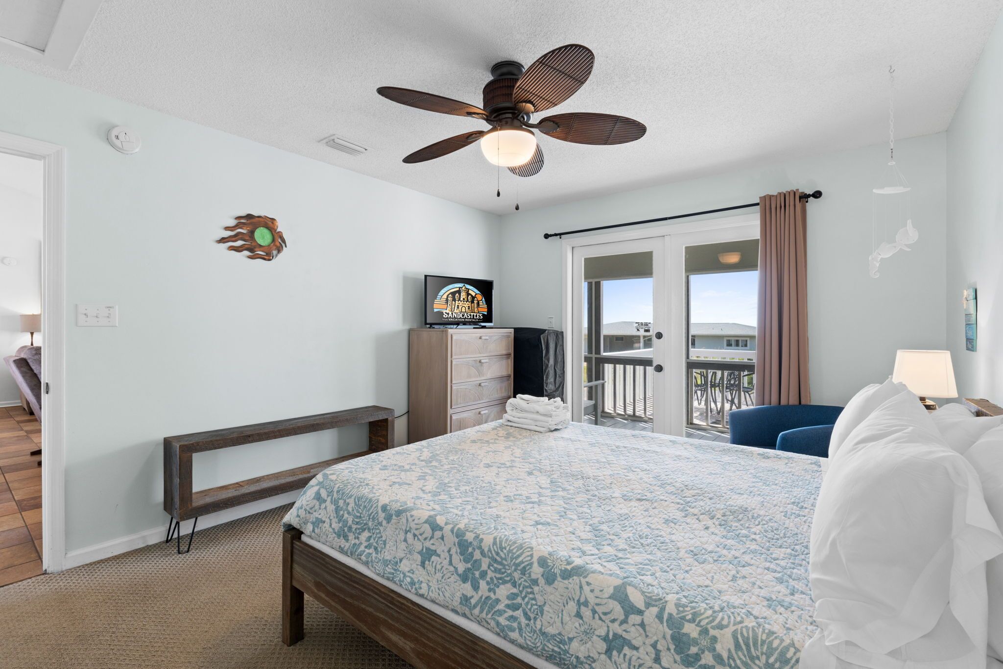 A bedroom with a bed, a ceiling fan, a TV, a bench, a desk, and a balcony with a view.