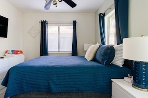 Un dormitorio con cama, mesita de noche, lámpara, ventilador de techo y ventana con cortinas.