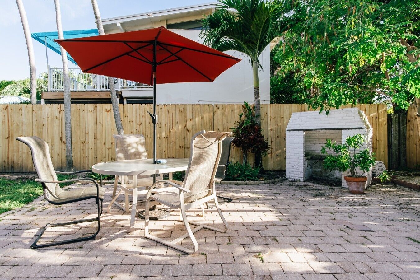 An outdoor patio with a round table, two chairs, a red umbrella, and a white brick fireplace.
