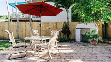 An outdoor patio with a round table, two chairs, a red umbrella, and a white brick fireplace.