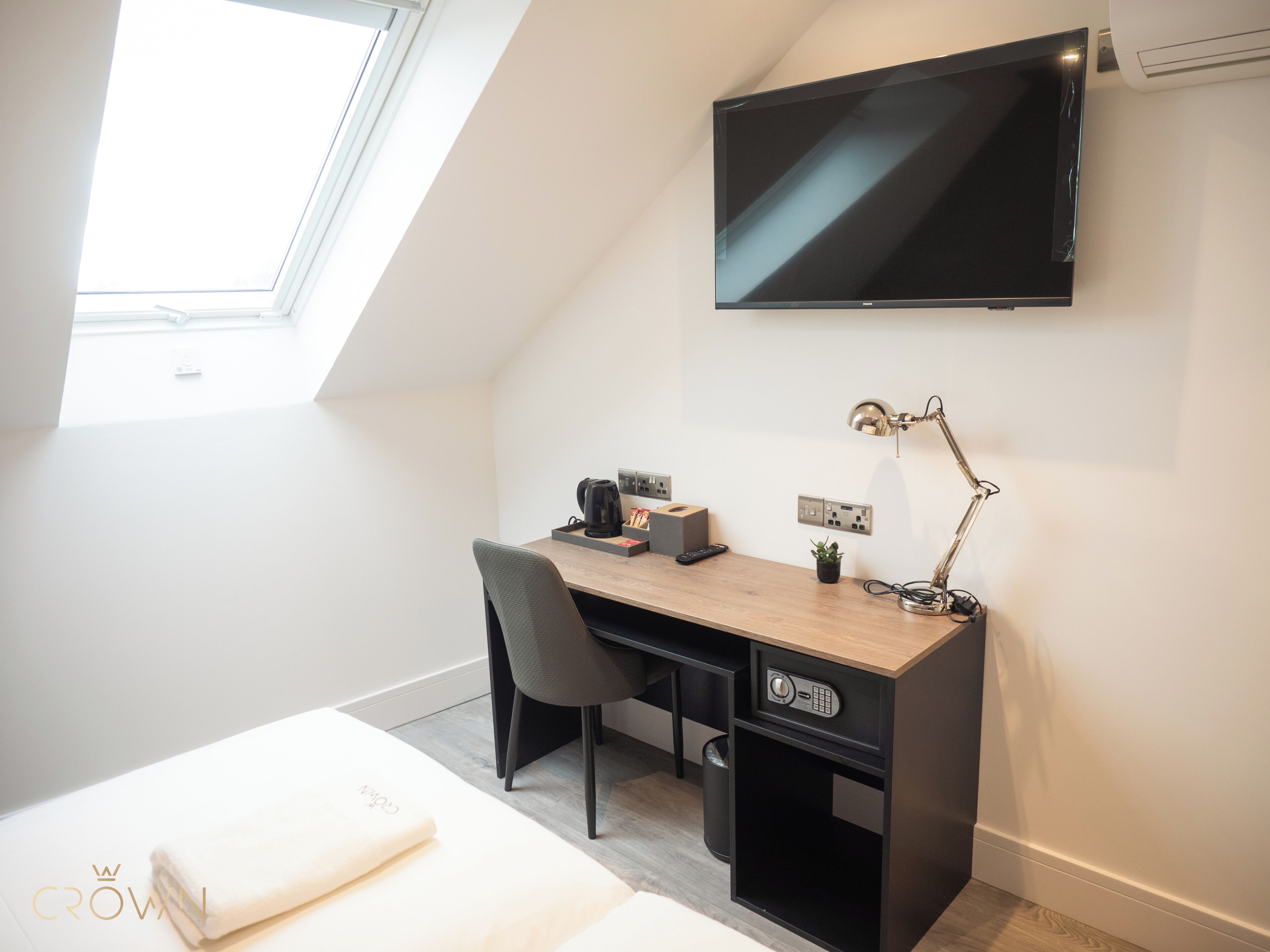 A modern bedroom with a skylight, a desk with a computer, a chair, a bed, and a TV mounted on the wall.