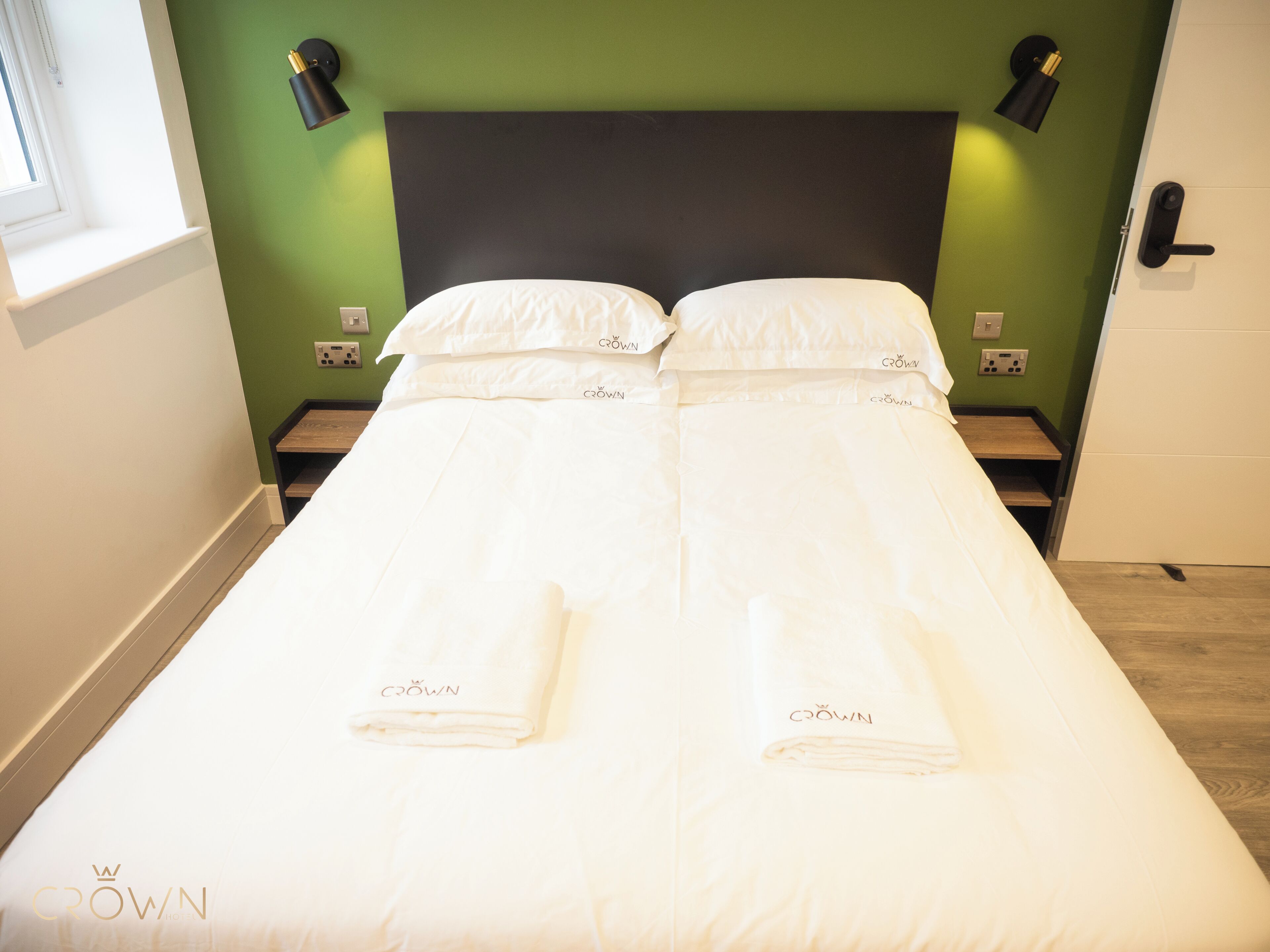 A double bed with white bedding and towels, two bedside tables, a black headboard, and green walls.