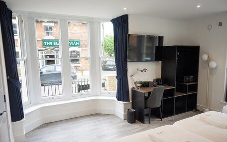 A hotel room with a desk, chair, and a bed. There is a window with curtains, a TV mounted on the wall, and a bookshelf.