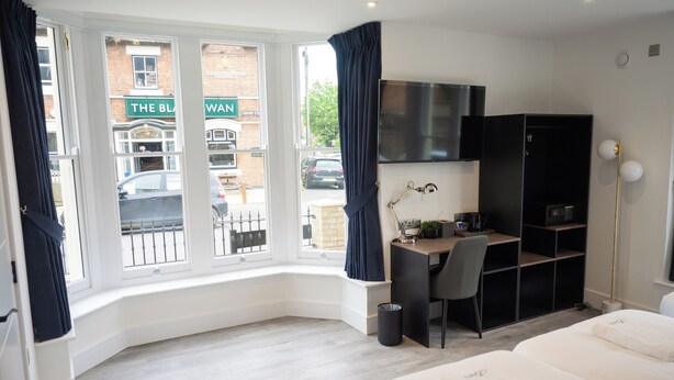 A hotel room with a desk, chair, and a bed. There is a window with curtains, a TV mounted on the wall, and a bookshelf. - Crown Hotel (Oxford)