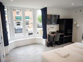 A hotel room with a desk, chair, and a bed. There is a window with curtains, a TV mounted on the wall, and a bookshelf.