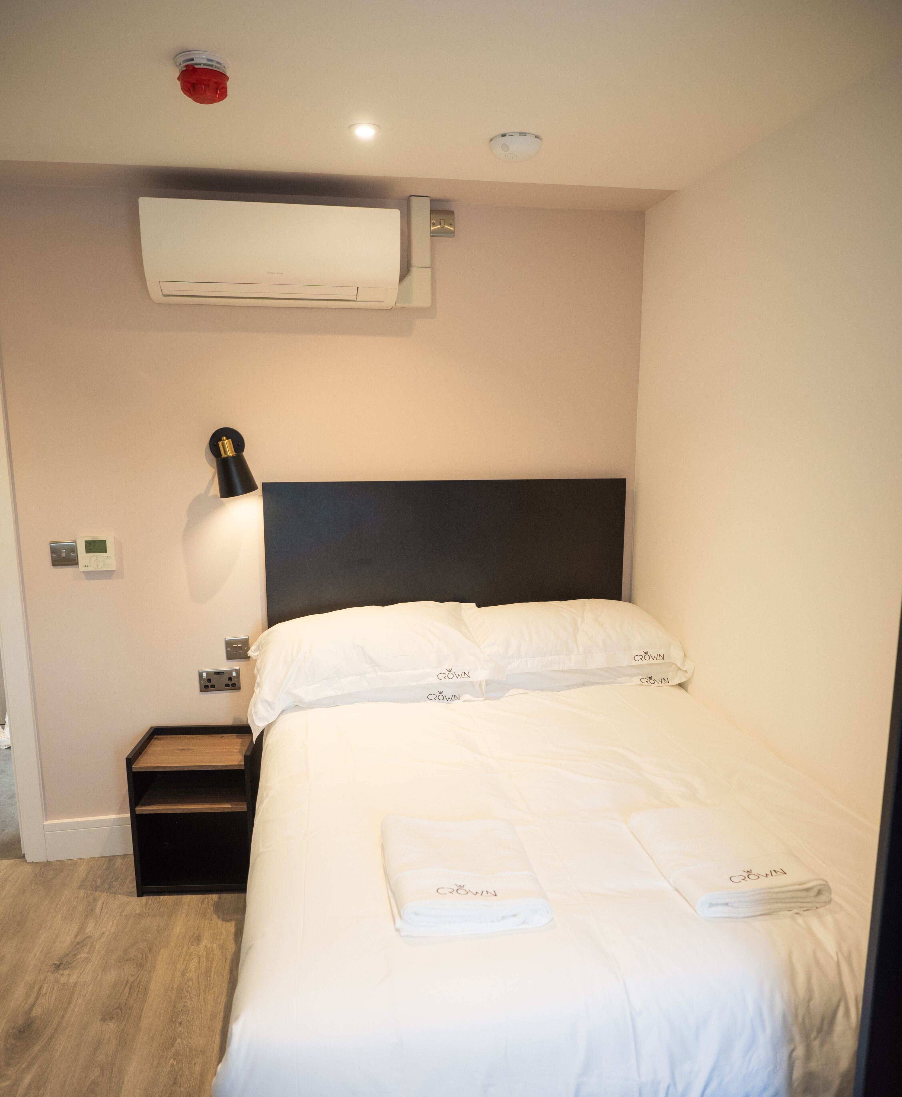 Hotellihuone, jossa on sänky, yöpöytä, seinään kiinnitetty ilmastointilaite, seinään kiinnitetty valaisin ja kattoon asennettu palovaroitin.