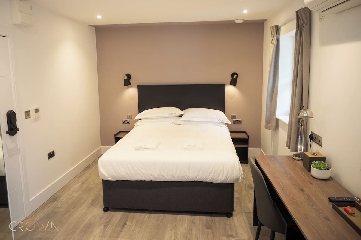 A hotel room with a bed, bedside tables, a desk with a chair, a potted plant, and a window with curtains.
