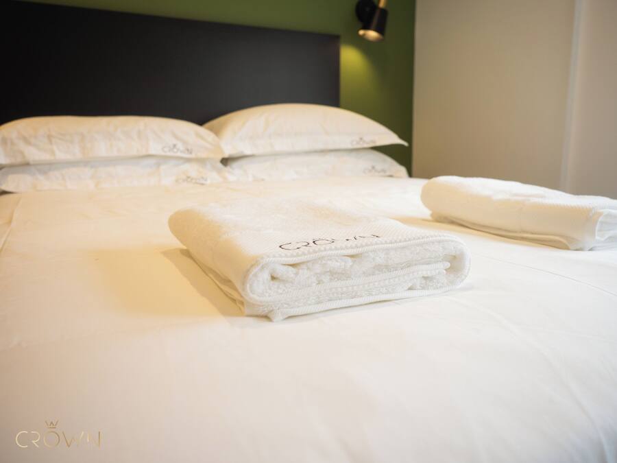 A neatly made bed with white linens and pillows, a folded towel on the bed, and a logo 'CROWN' on the headboard.
