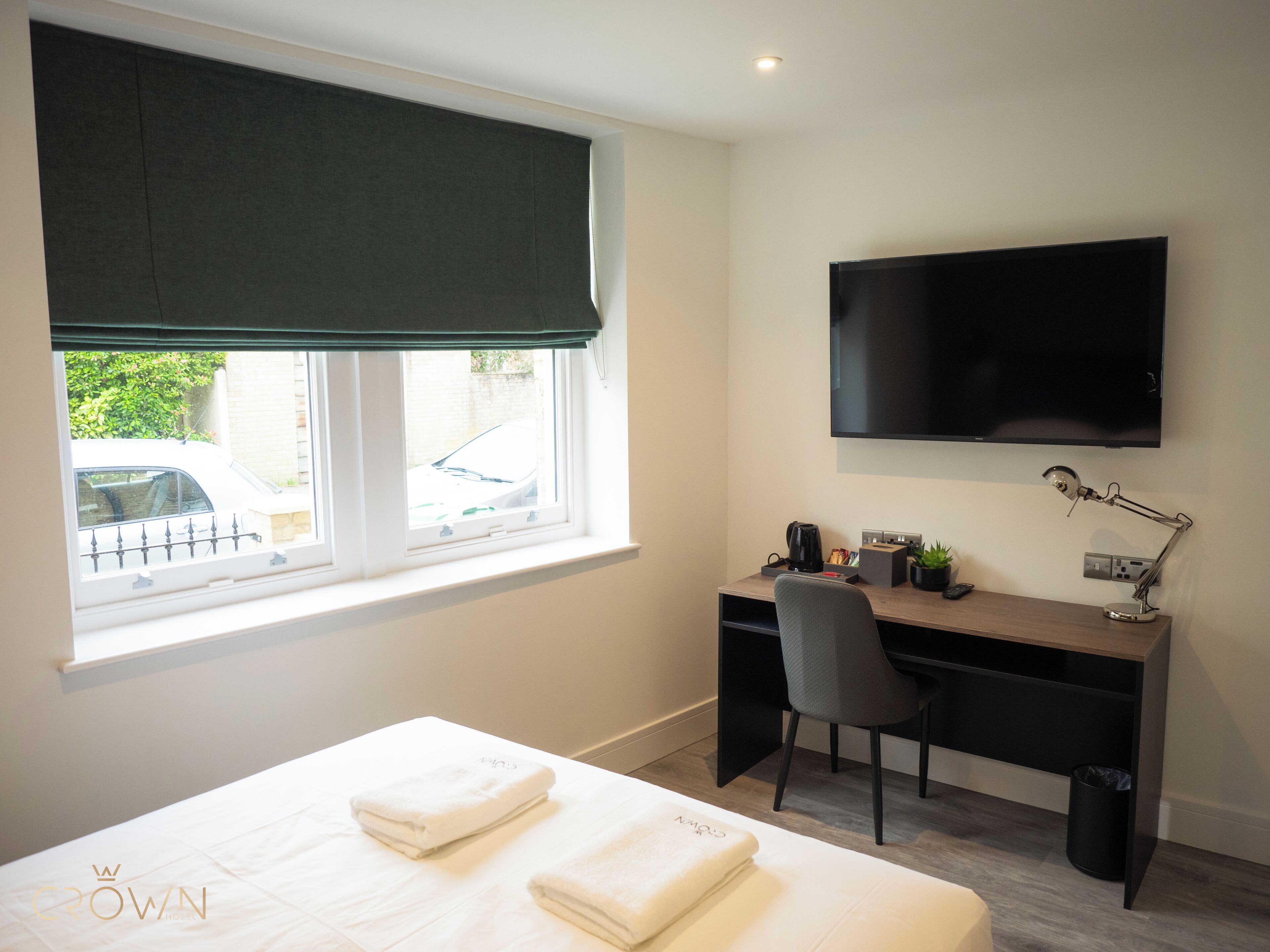 A hotel room with a bed, a desk, a chair, a television, and a window with a view of a car and greenery outside.