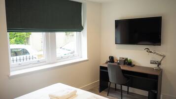 A hotel room with a bed, a desk, a chair, a television, and a window with a view of a car and greenery outside.