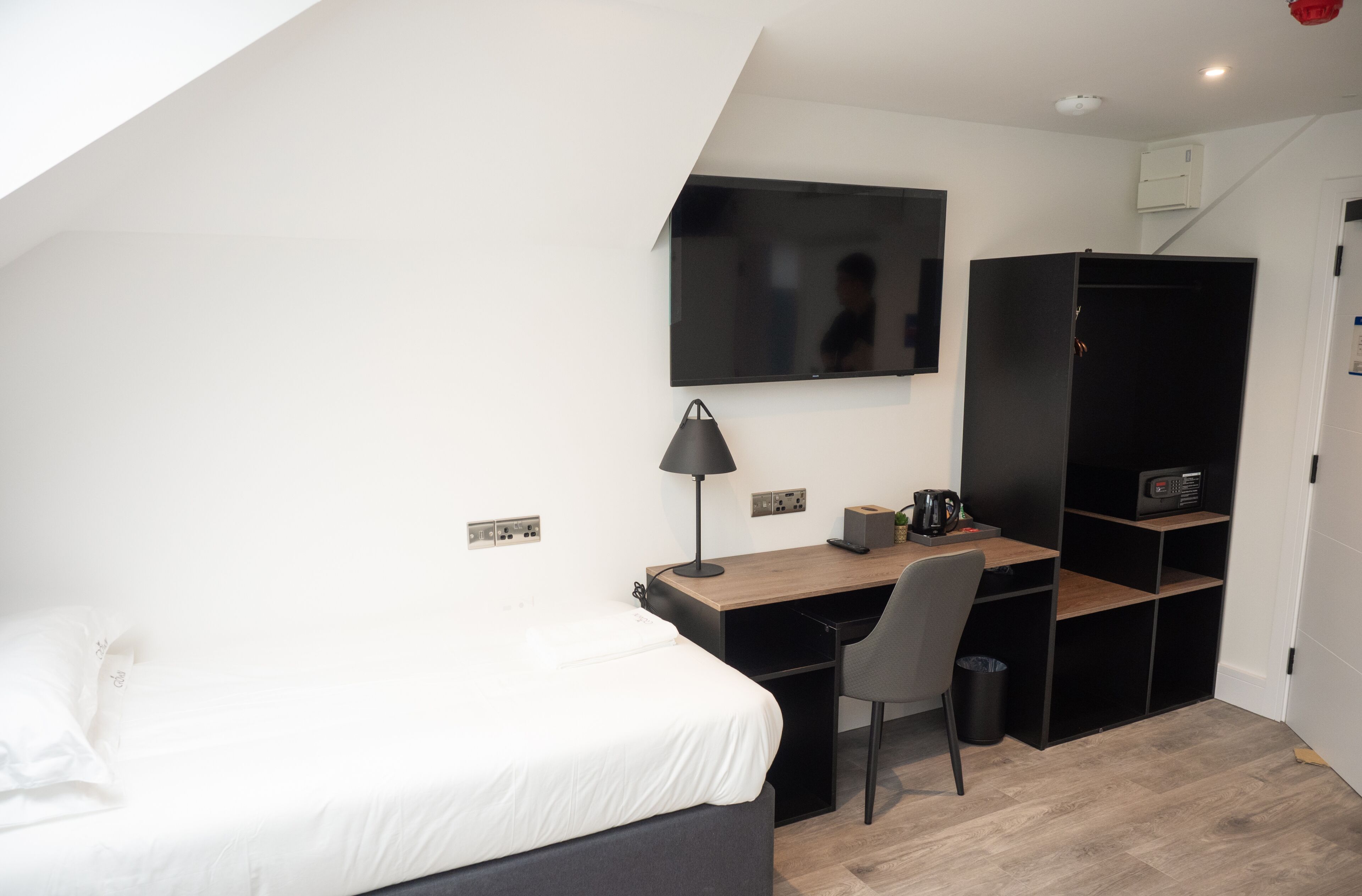 Habitación de hotel pequeña con cama, escritorio, silla, televisor montado en la pared y armario.