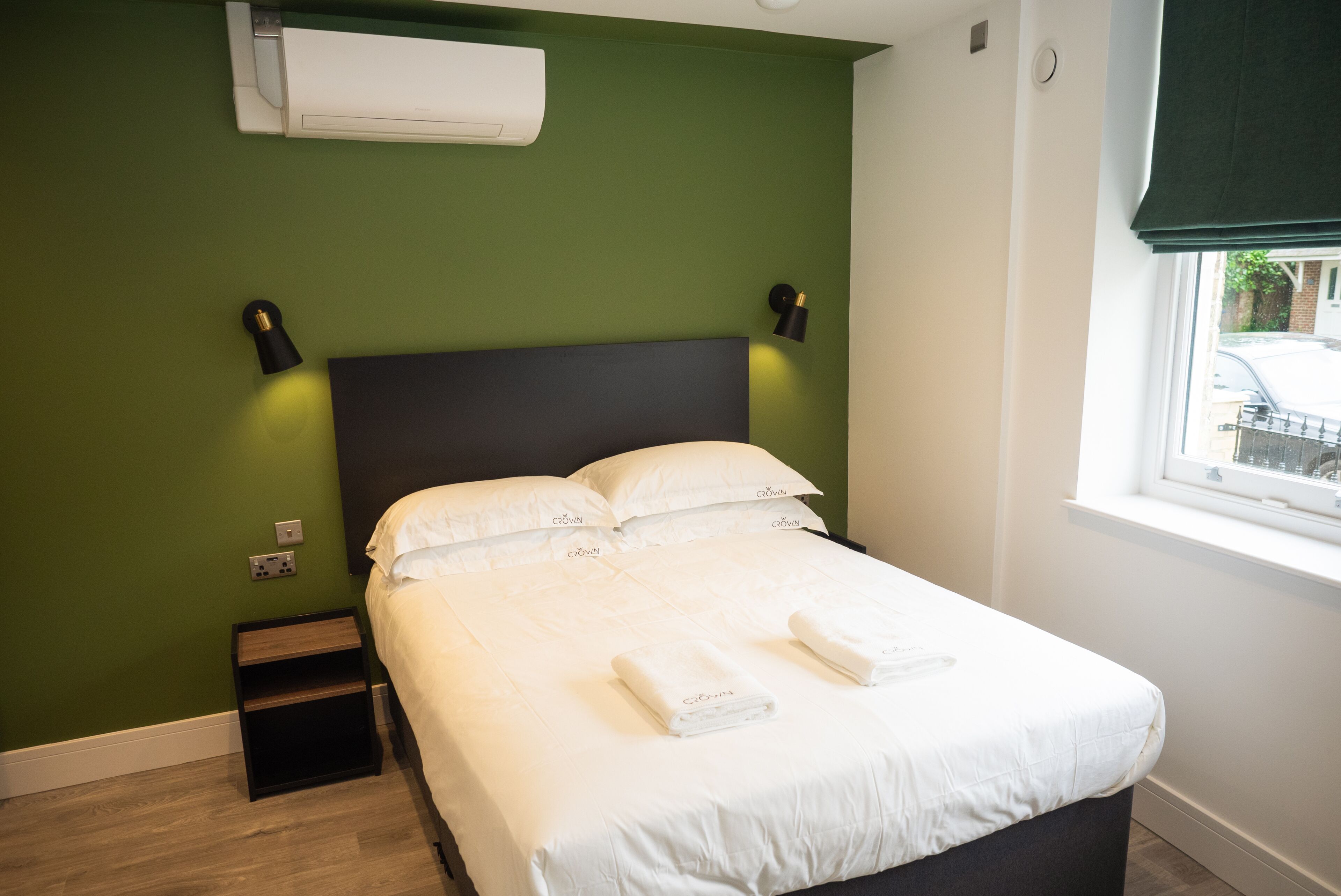 Una cama bien hecha con ropa de cama y toallas blancas, un cabecero oscuro, una mesita de noche y un aire acondicionado de pared.