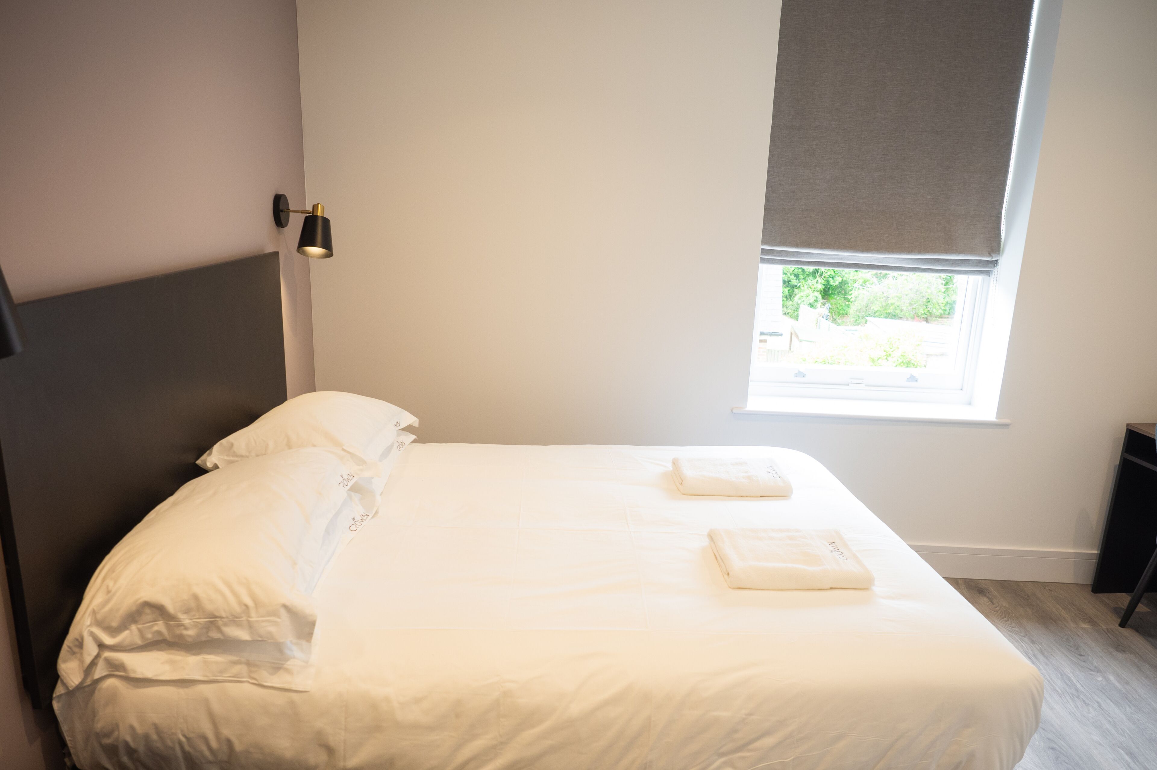 A neatly made bed with pillows and a folded towel, a bedside lamp, and a window with a view of greenery.